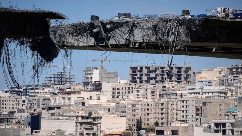 A severely damaged concrete structure with exposed rebar hangs over a dense cityscape of multi-story buildings under a clear sky.