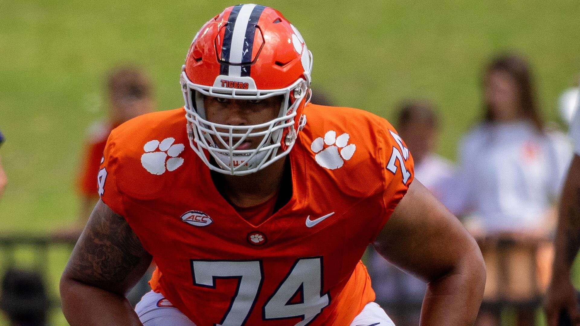 A Clemson Tigers football player wearing an orange #74 jersey and helmet looks forward in a ready stance on the field.
