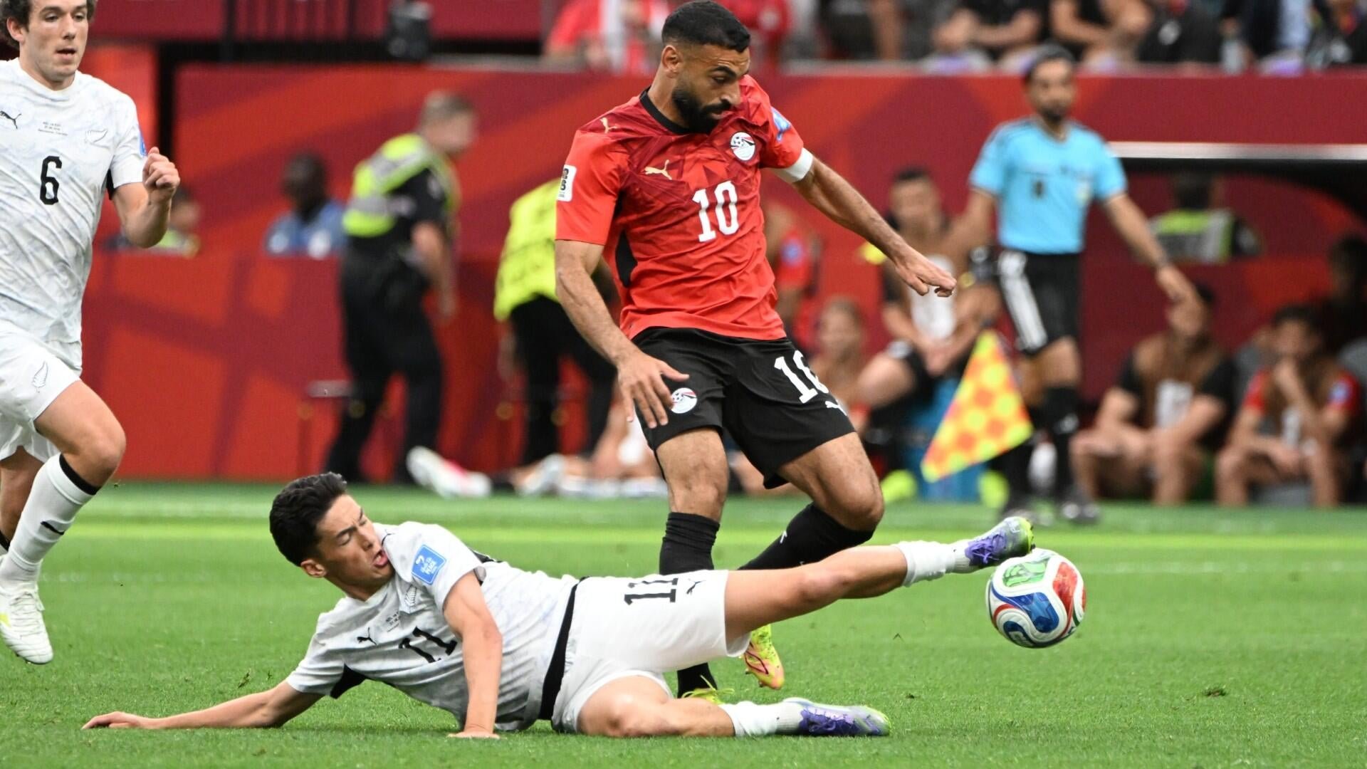 A soccer player in a red jersey dribbles the ball past a sliding defender in white during a match, with a referee and spectators visible in the background.