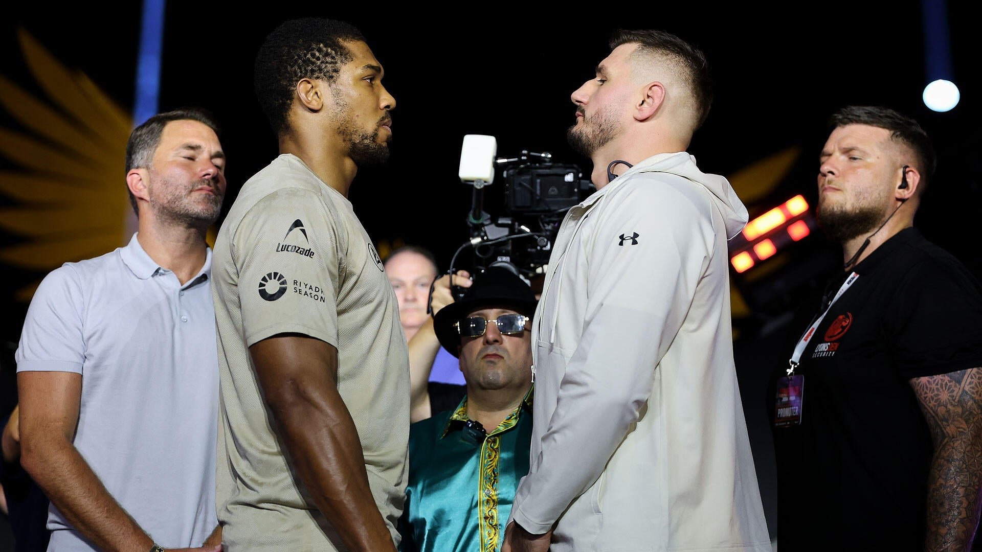 Two men face each other in a stare-down during a promotional event, surrounded by camera crew and staff, with one wearing a Lucozade/Riyadh Season shirt and the other a white hoodie.