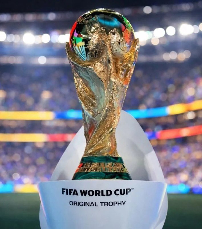 A golden FIFA World Cup trophy sits on a white stand reading "FIFA WORLD CUP ORIGINAL TROPHY," with a blurred stadium and crowd visible in the background.