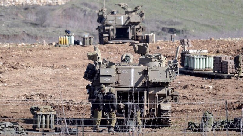 Two armored military vehicles and soldiers in helmets are positioned in a rocky, barren landscape behind barbed wire, with artillery shells visible nearby.