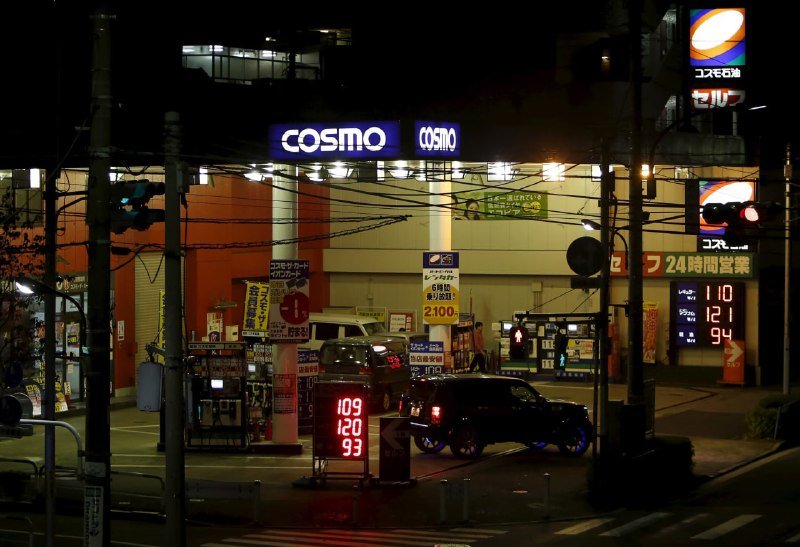A nighttime gas station with illuminated "COSMO" signage, fuel price displays showing 109, 120, and 93, and a black SUV with glowing blue undercarriage lights parked at the pumps.