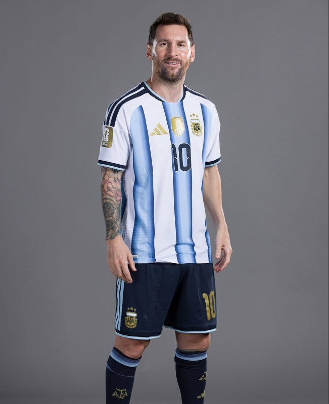 A bearded man with arm tattoos poses in a light blue and white striped soccer jersey bearing the number 10 and a team crest, against a gray studio backdrop.