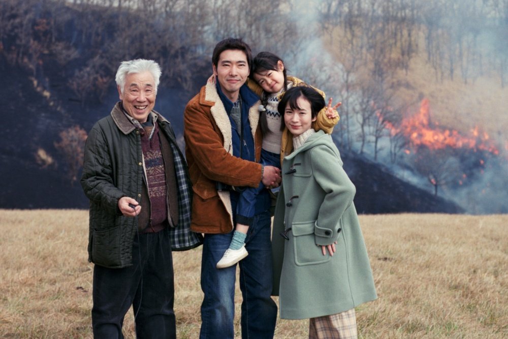 Four people, including a child, pose smiling in a field with a burning hillside and smoke visible in the background.