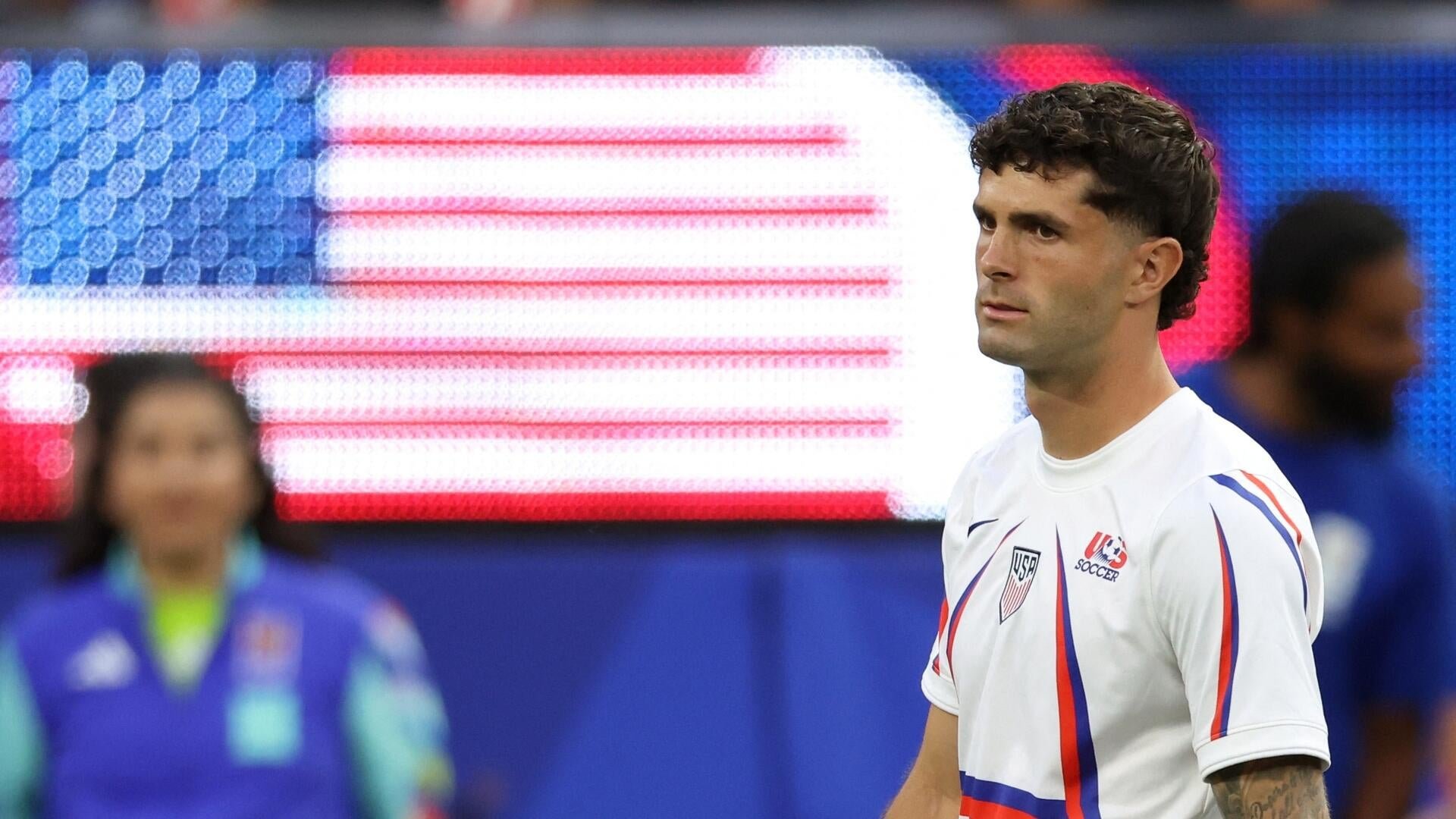 A US Soccer player in a white jersey stands on the field with an American flag displayed on a screen behind him.