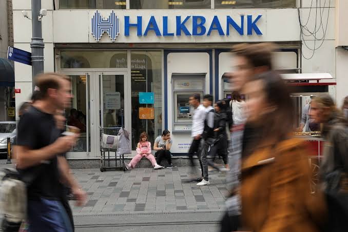 Pedestrians walk past a Halkbank branch with an ATM, where two people sit on the sidewalk near the entrance.