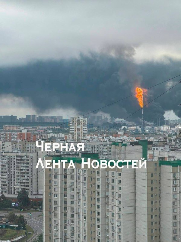 A large plume of black smoke rises above a city skyline as a tall flame burns from an industrial structure, with the text "ЧЕРНАЯ ЛЕНТА НОВОСТЕЙ" overlaid.