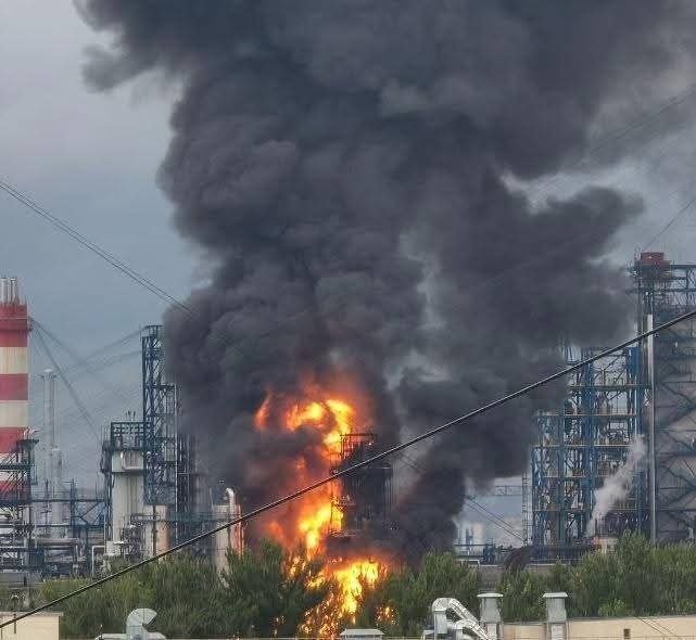 Thick black smoke and bright orange flames rise from an industrial facility with tall structures and piping, visible behind a row of green trees.