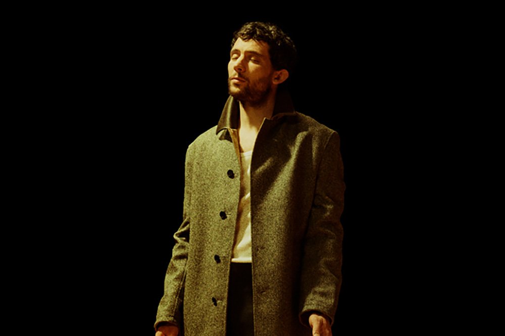 A bearded man in a gray wool overcoat stands against a black background with his eyes closed, illuminated by warm side lighting.
