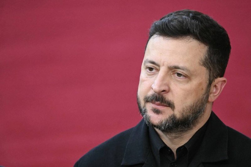 A bearded man in a dark jacket and shirt looks off to the side against a solid pinkish-red background.