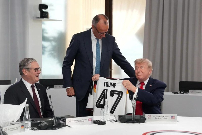 At a G7 summit meeting, three men in suits examine a white "TRUMP 47" jersey, with one man seated smiling and another holding the jersey up.