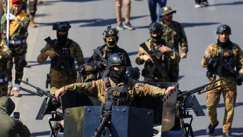 Armed personnel in camouflage uniforms, helmets, and face coverings stand with rifles mounted on a vehicle equipped with metal shields during a street procession.