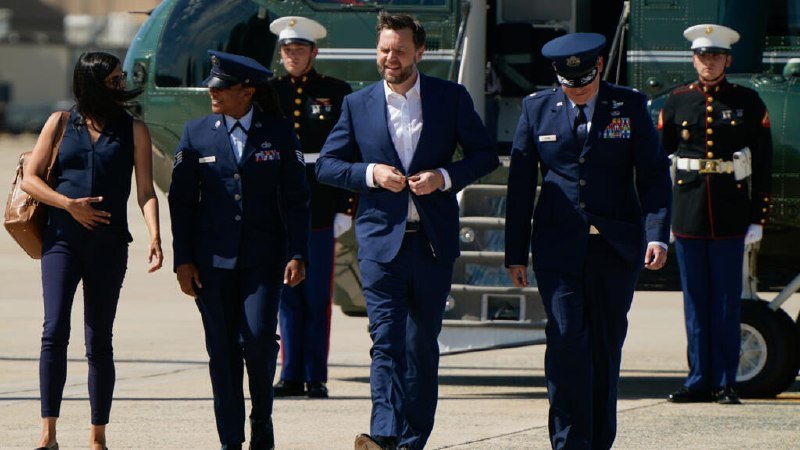A man in a blue suit walks alongside military personnel in dress uniforms and a woman on a tarmac, with a Marine Corps helicopter visible in the background.