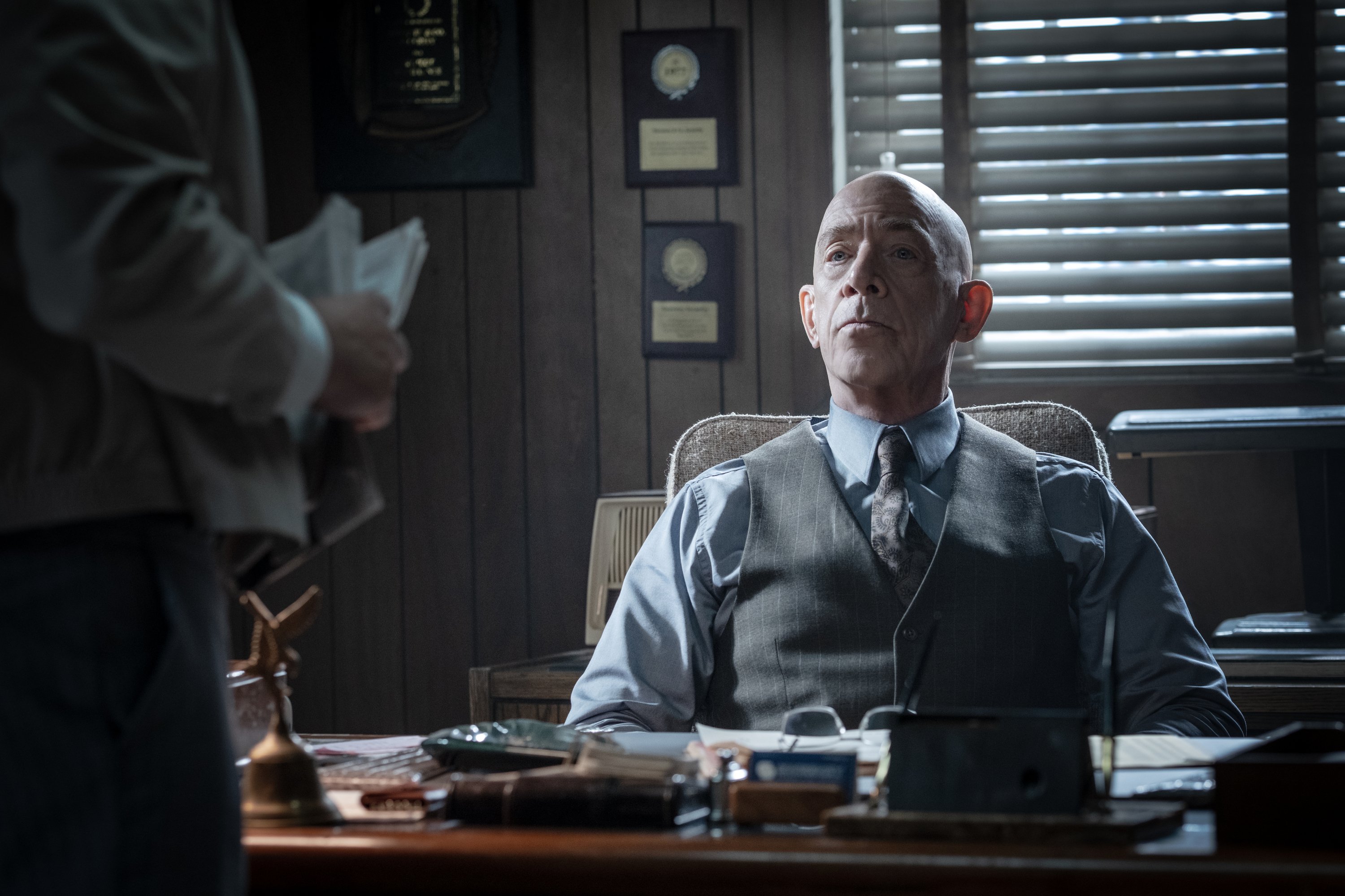 A bald man in a vest and tie sits at a cluttered desk in a dim office, looking up toward a standing person holding papers, with framed plaques on the wood-paneled wall behind him.