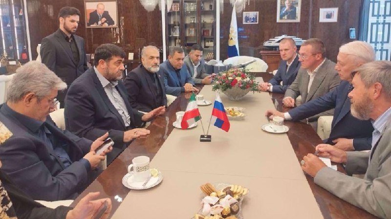 Two delegations sit across a conference table adorned with Iranian and Russian flags, floral centerpiece, teacups, and sweets, engaged in discussion.