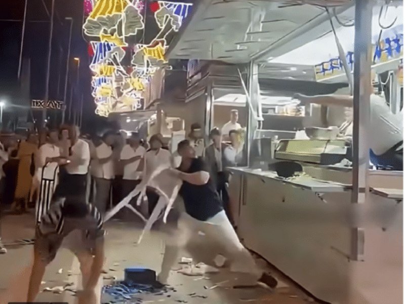 Two women wrestle over a white plastic chair at night near a food truck, with scattered debris on the ground and onlookers watching.