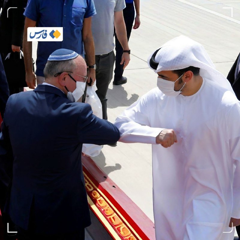 Two masked men, one wearing a kippah and dark suit and the other in traditional white Arab attire, bump elbows over a red carpet.