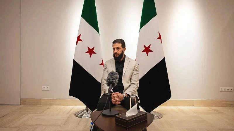 A bearded man in a light jacket sits before a microphone flanked by two green, white, and black flags with red stars, in a sparse room with a small table.