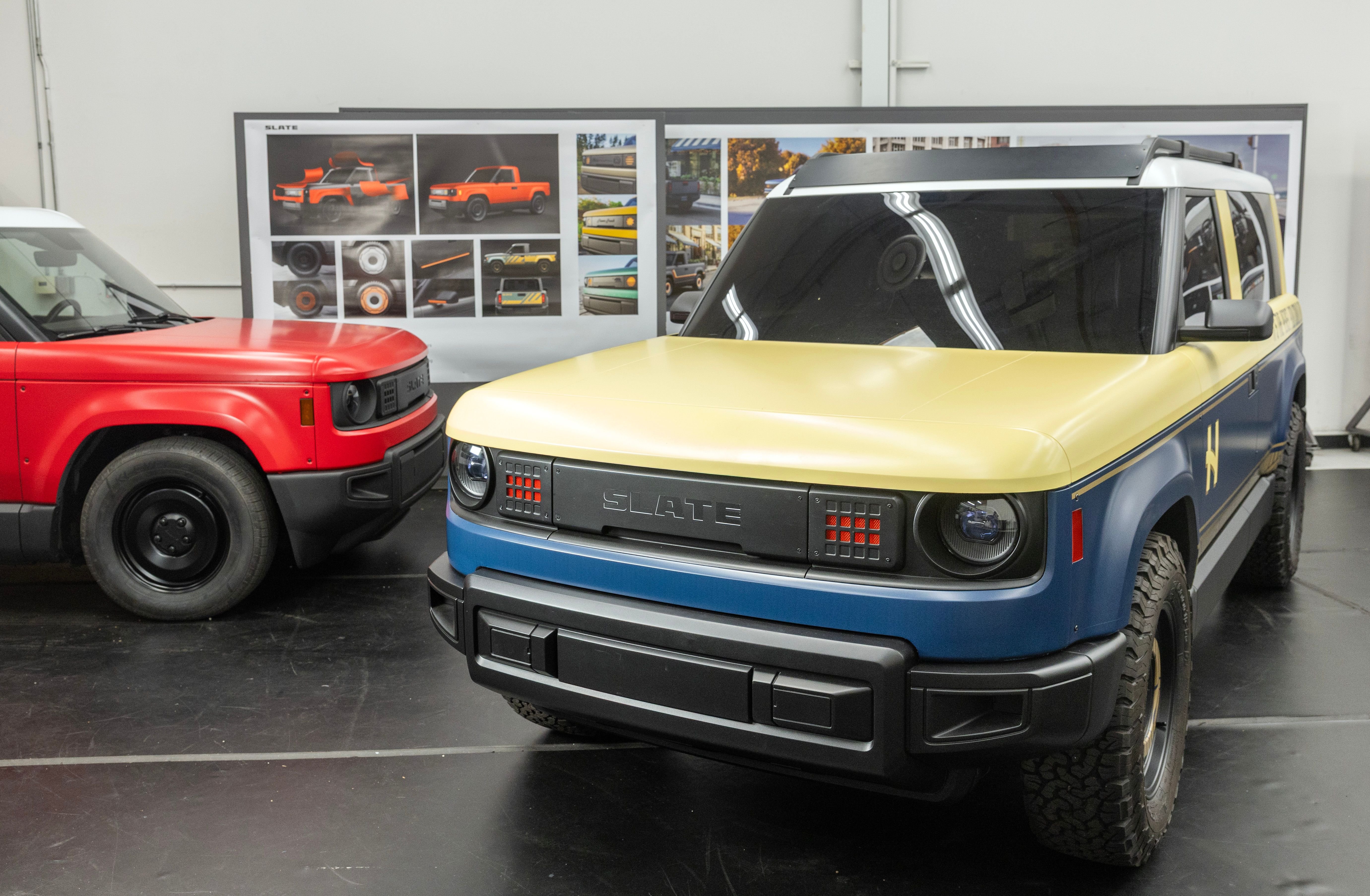 Two compact SUVs branded "SLATE" — one yellow and blue, one red — are displayed indoors on a black platform in front of design presentation boards.