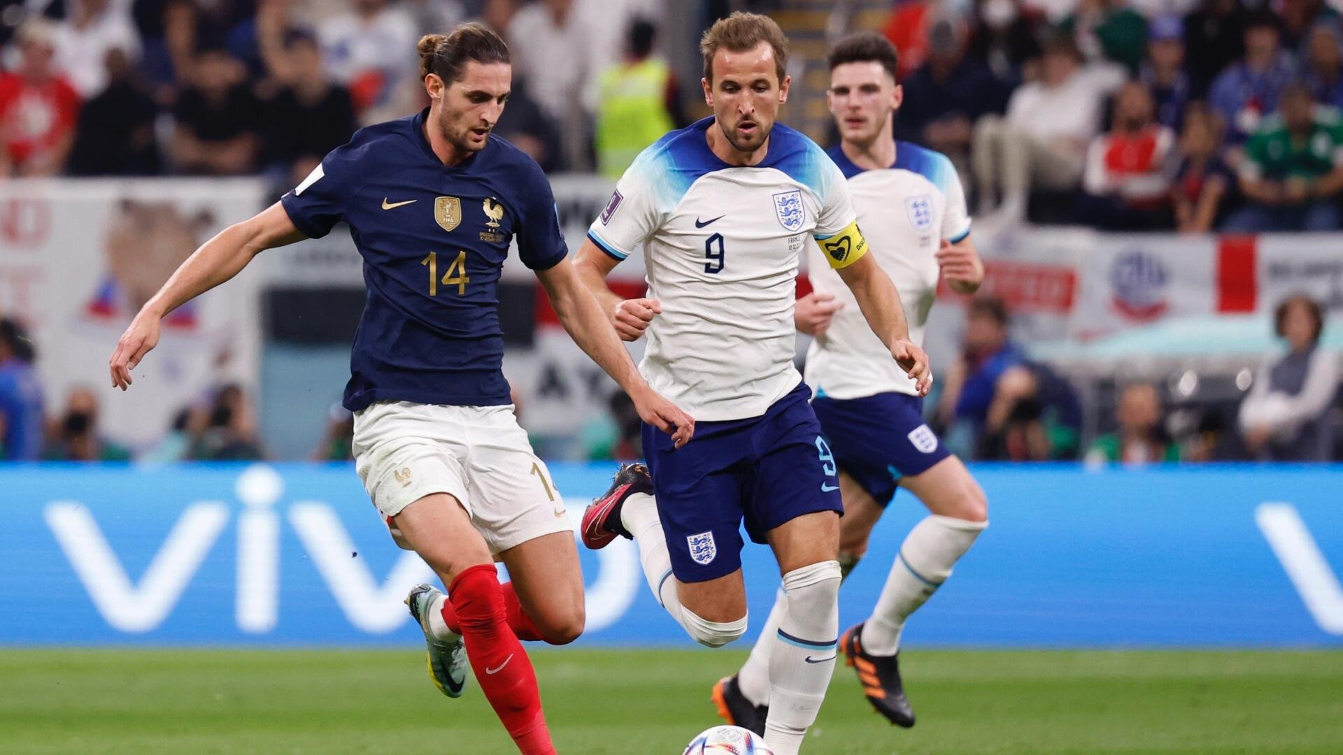A soccer player in a white England jersey numbered 9 dribbles the ball while being pursued by a player in a dark blue France jersey numbered 14, with another England player and spectators visible in the background.