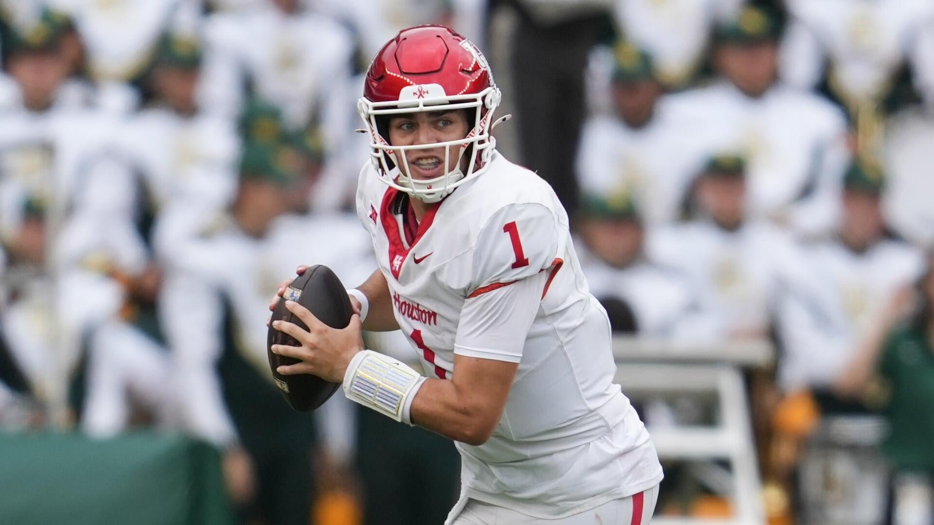 A football player in a white Houston jersey (#1) and red helmet holds a football while looking to pass during a game, with opposing players in green and white visible in the background.