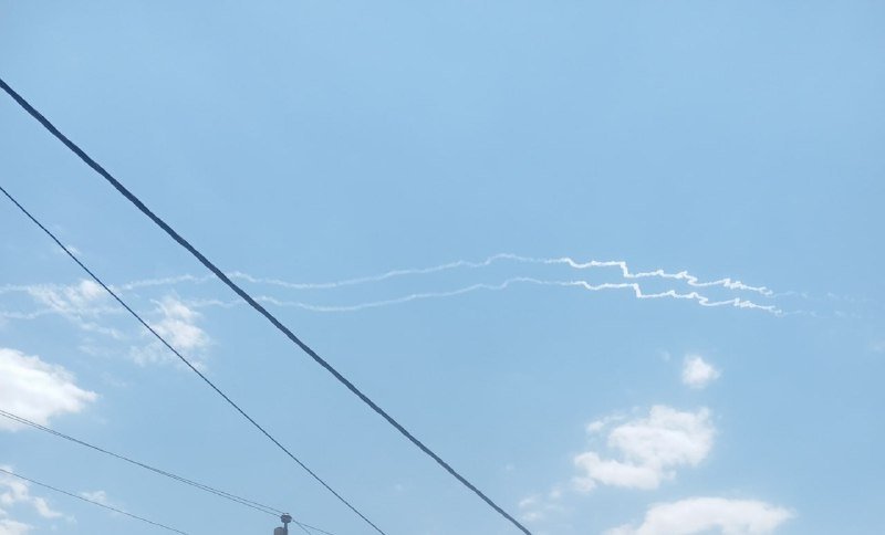 White contrails stretch across a blue sky with scattered clouds, while power lines cut diagonally through the foreground.