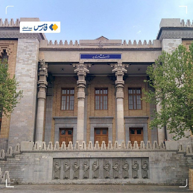 A grand stone building features tall ornate columns, multiple arched doorways, a blue Persian-script sign above the entrance, and a frieze of carved figures lining the lower wall.