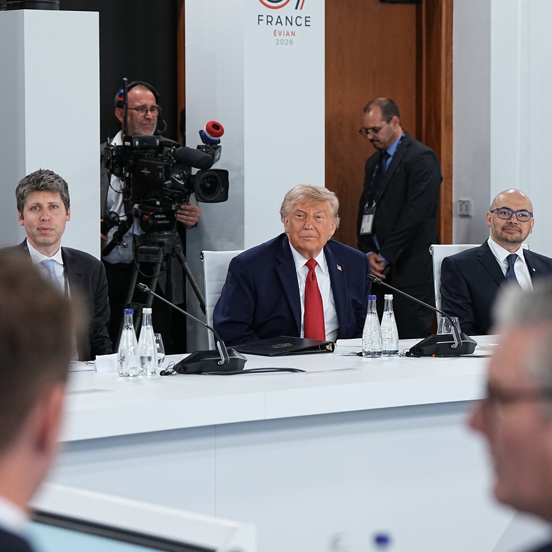 A man in a dark suit and red tie sits at a conference table with microphones and water bottles, flanked by other officials, while a cameraman films in front of a "FRANCE ÉVIAN 2026" backdrop.