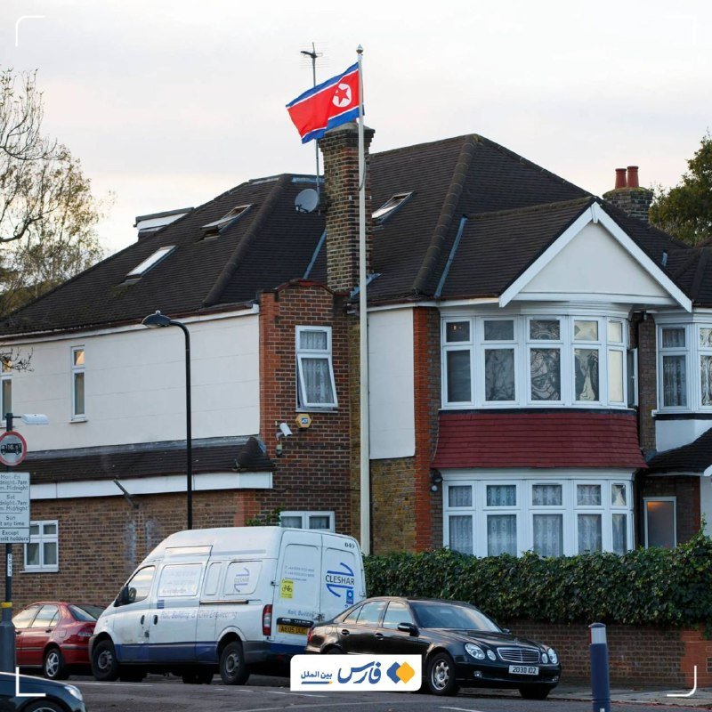 The North Korean flag flies from a flagpole in front of a two-story brick house, with a white van and black car parked on the street below.