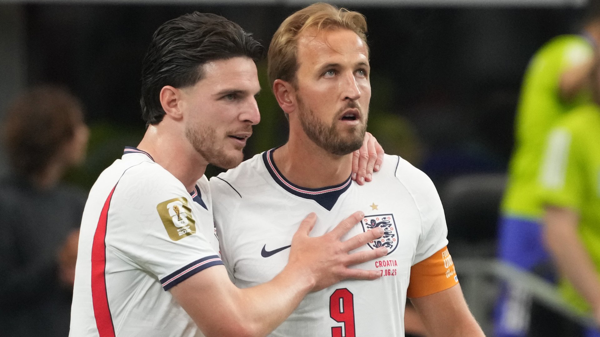 Two soccer players in white England jerseys embrace, with the player wearing number 9 and a captain's armband placing his hand on his chest.