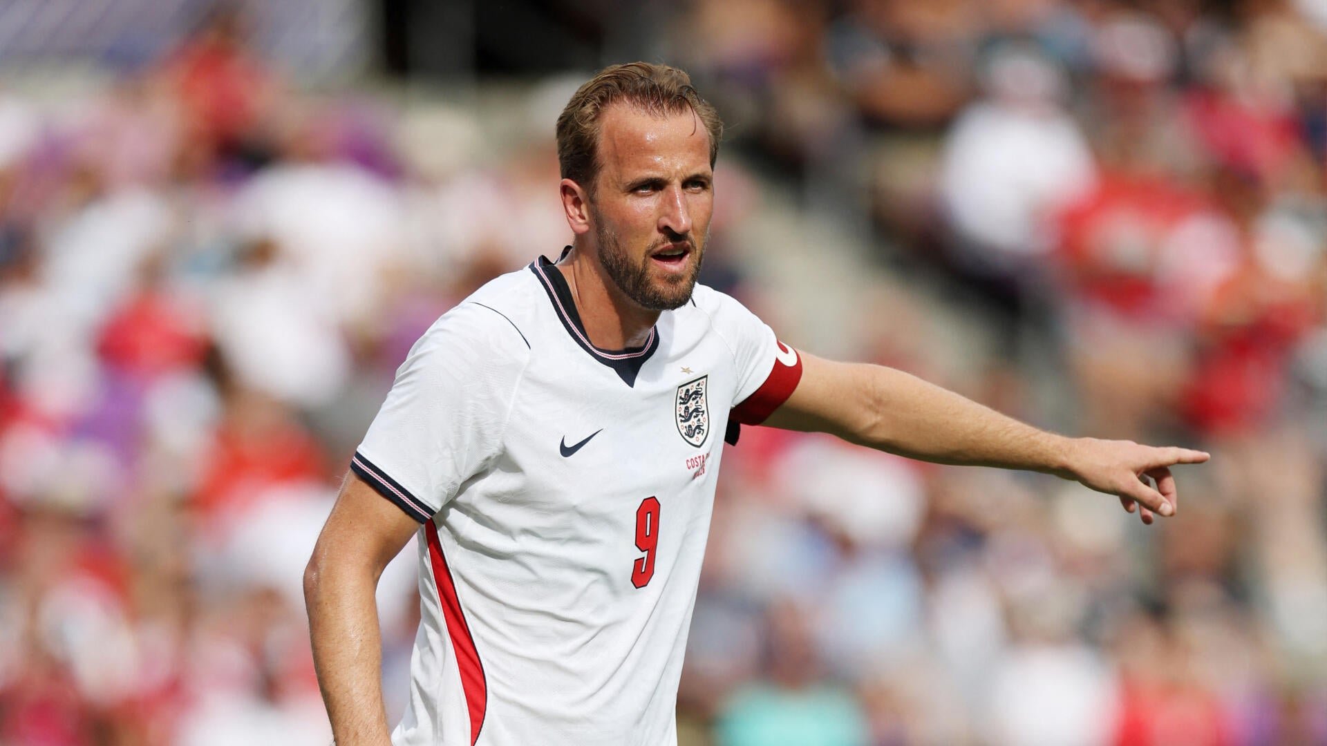 A bearded soccer player in a white England jersey with the number 9 and a red captain's armband points forward on the pitch during a daytime match.
