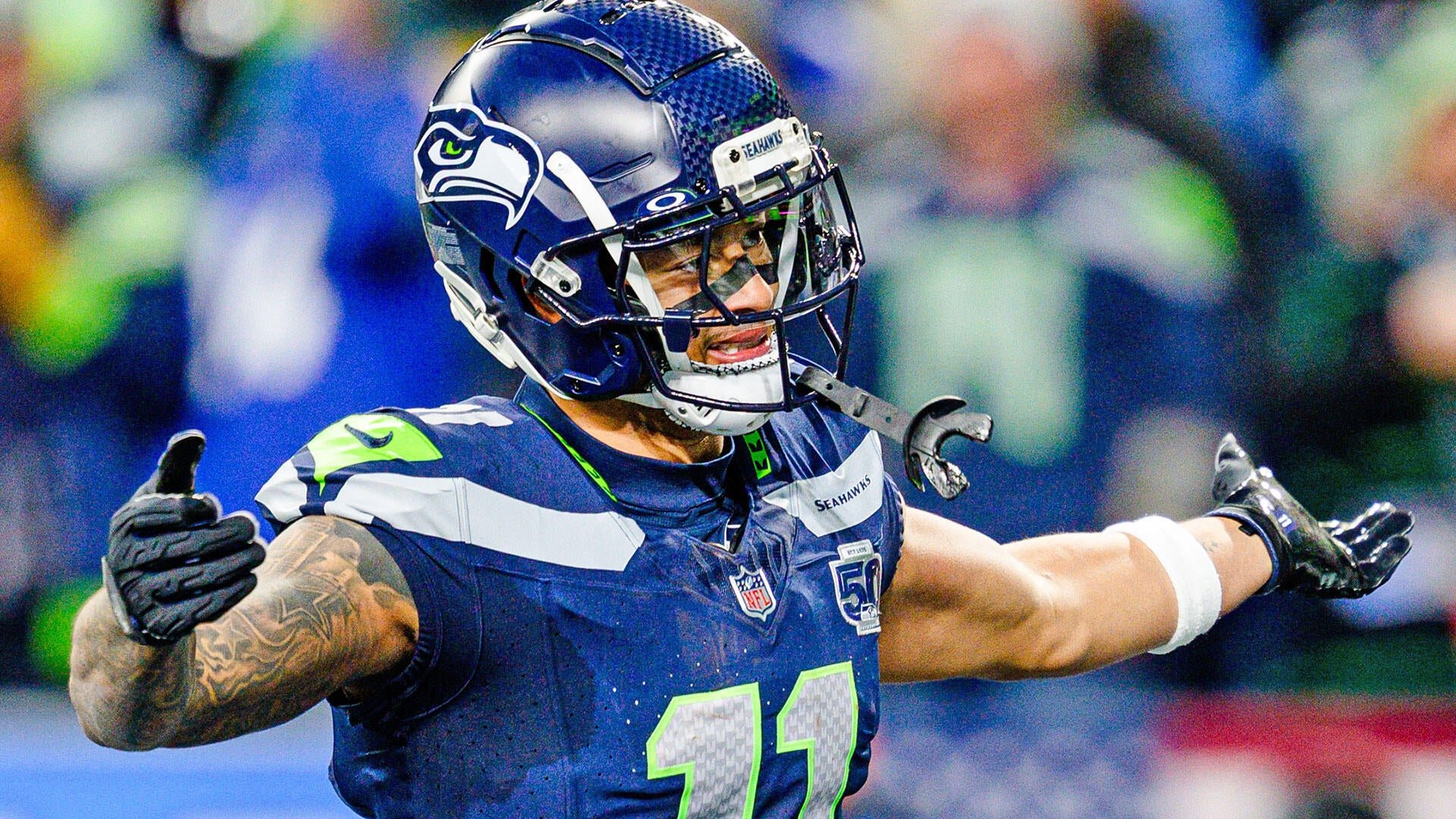 A Seattle Seahawks football player in a navy blue uniform and helmet celebrates with arms outstretched on the field during a game.