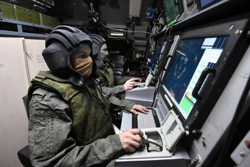 Two helmeted, masked personnel in camouflage uniforms operate computer consoles and control devices inside a cramped, equipment-filled mobile command vehicle.