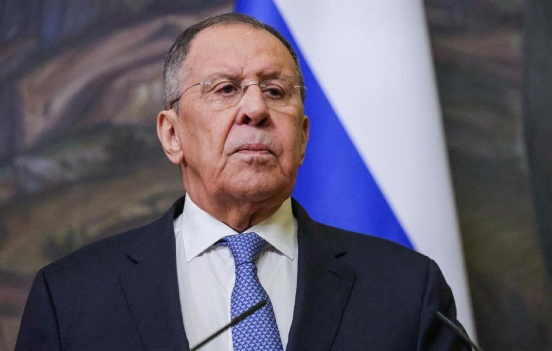 An older man in a dark suit, white shirt, and blue patterned tie stands before a Russian flag, appearing to speak at a podium.