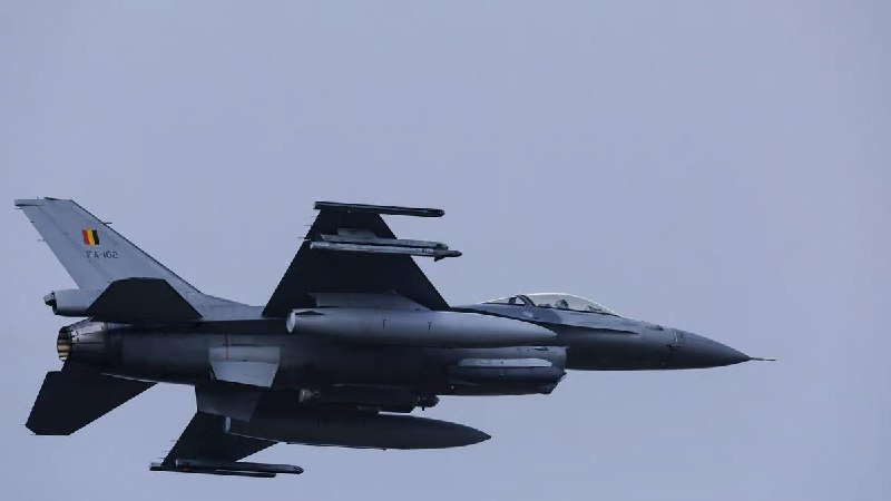 A gray F-16 fighter jet with tail markings "FA-60" and a black-yellow-red flag flies against a clear sky, carrying external fuel tanks and weapons.