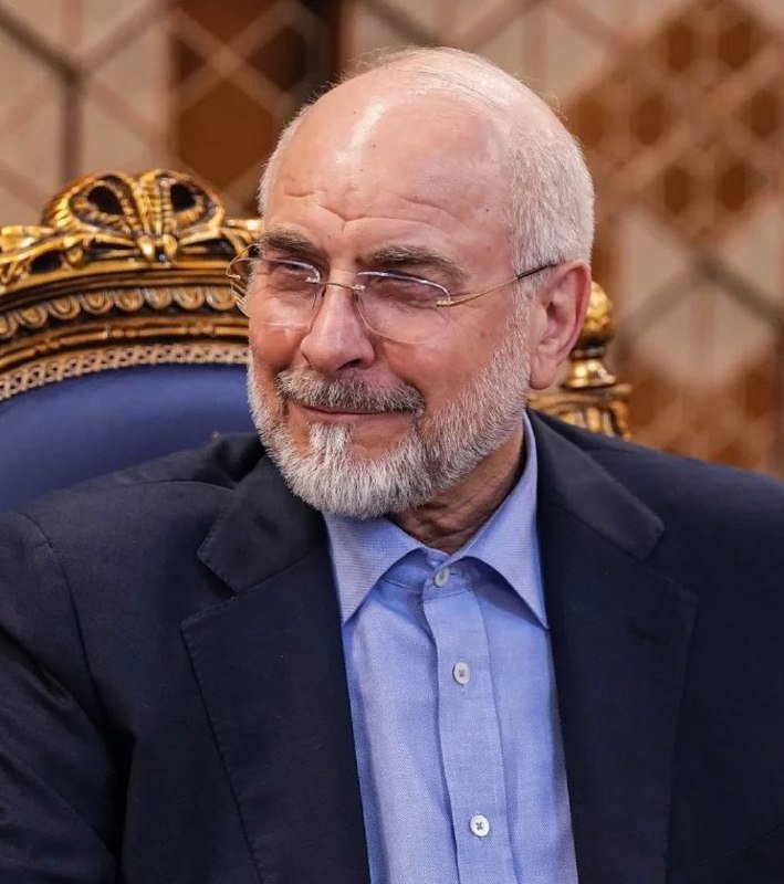 A bearded, balding man with glasses smiles while seated in an ornate chair with gold detailing, wearing a dark blazer over a blue shirt.