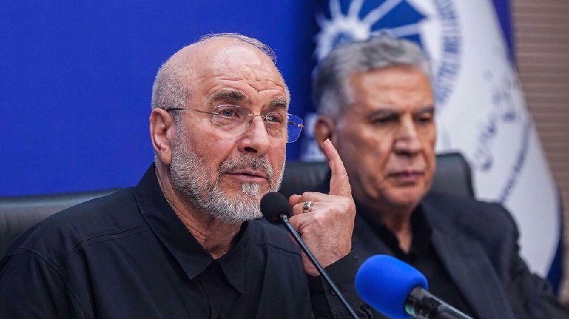 Two bearded men in dark shirts sit before microphones at a press conference, with one gesturing with his index finger raised beside a partial blue flag emblem.