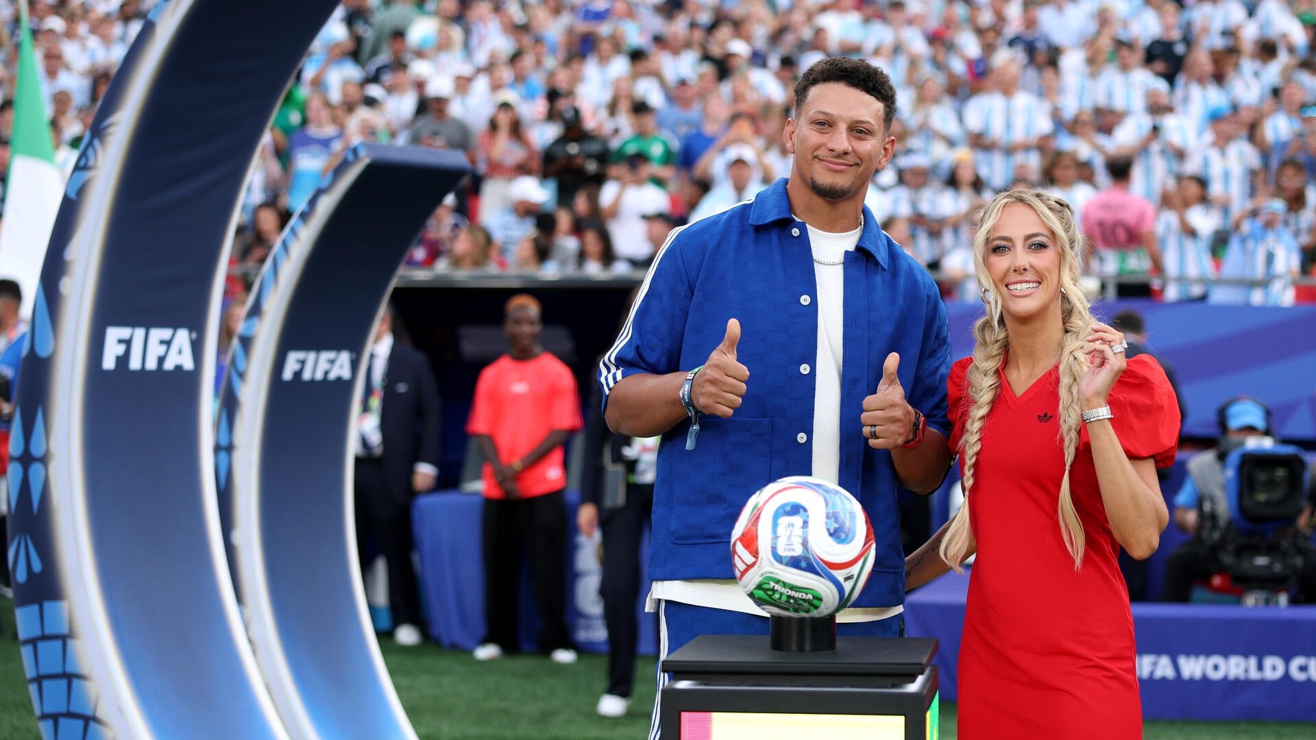A man in a blue jacket and a woman in a red dress give thumbs-up beside a trophy soccer ball on a pedestal marked "FIFA," with a crowded stadium behind them.