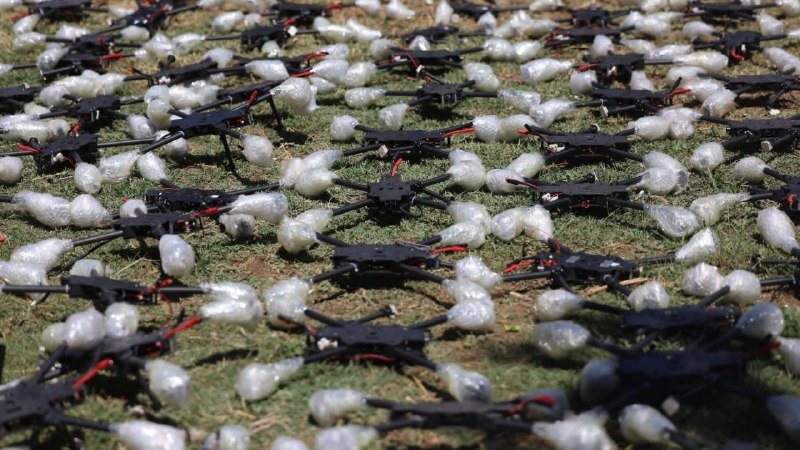Numerous black quadcopter drones and white bubble-wrapped packages are scattered across a grassy field.