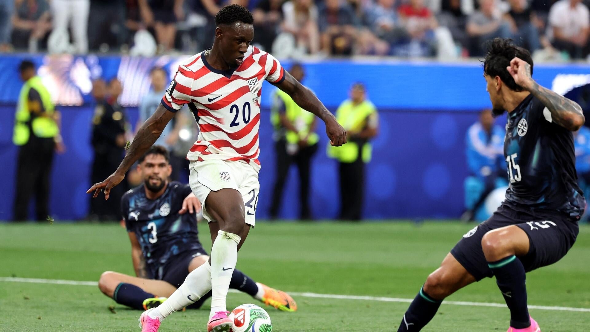 A USA soccer player in a red, white, and blue striped jersey numbered 20 dribbles the ball while two opposing players in dark jerseys attempt to defend during a match.