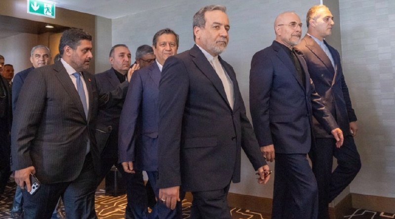 A group of men in dark suits walk together through a hotel corridor with patterned carpet and an illuminated exit sign overhead.