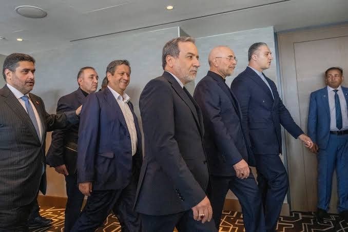 A group of seven men in dark suits walk together through a modern interior hallway with light walls and patterned carpeting.