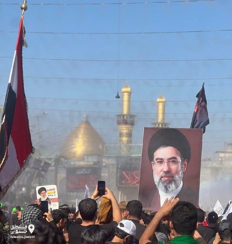 A crowd holds up a large portrait of a bearded cleric in black robes and turban, with a golden-domed shrine and minarets visible behind them.