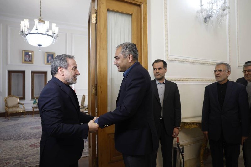 Two men in dark suits shake hands in a chandelier-lit formal room while three other men stand nearby observing.