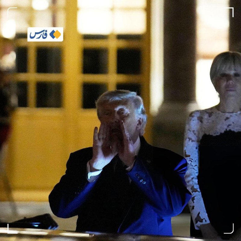 A man in a dark suit stands with both hands cupped near his mouth at night, while a woman in a dark dress stands behind him near a yellow building.
