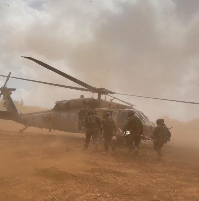 Five helmeted soldiers in camouflage uniforms walk toward a grounded military helicopter as thick dust swirls around them under an overcast sky.