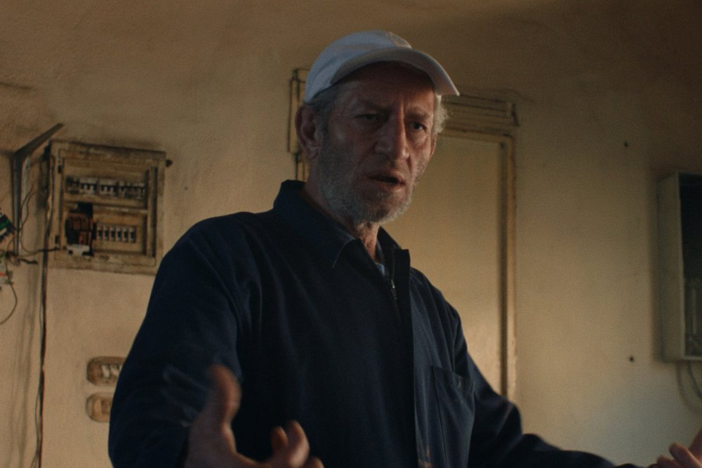 A bearded man wearing a light blue cap and dark jacket speaks with a concerned expression inside a room with an exposed electrical panel.
