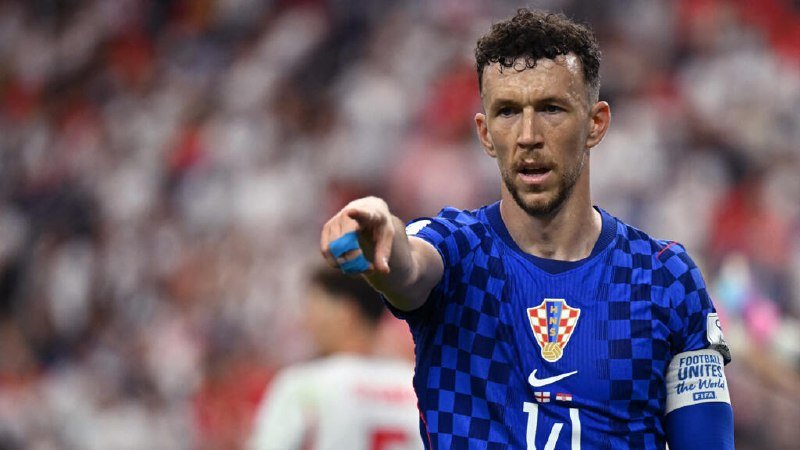 A soccer player wearing a blue checkered Croatia jersey points forward during a match, with a blurred stadium crowd in the background.