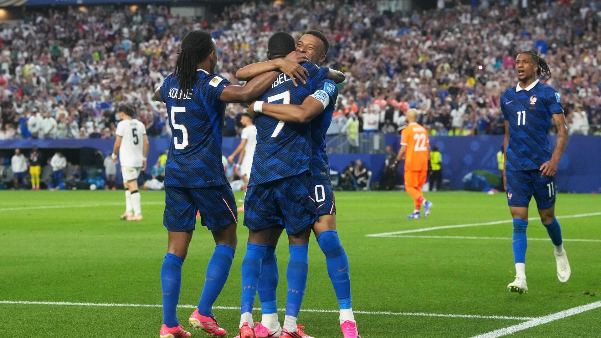 Soccer players in blue jerseys celebrate together on a stadium pitch while opposing players in white and an orange-uniformed goalkeeper stand nearby before a packed crowd.